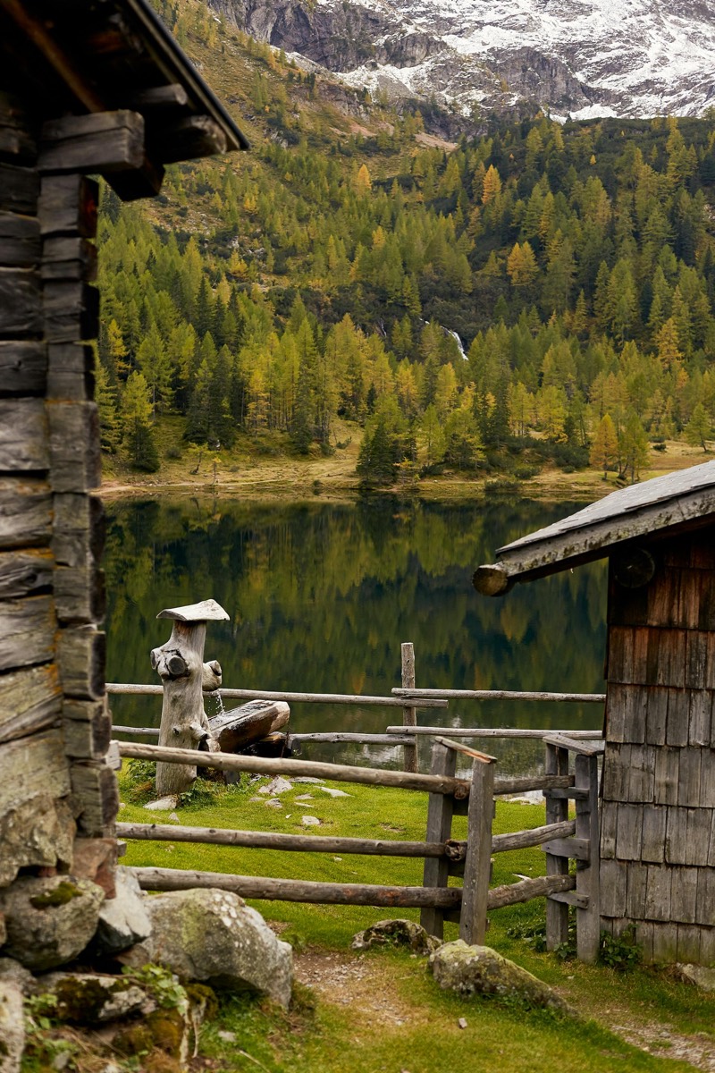 A wooden cabin beside a calm alpine lake with mountains behind